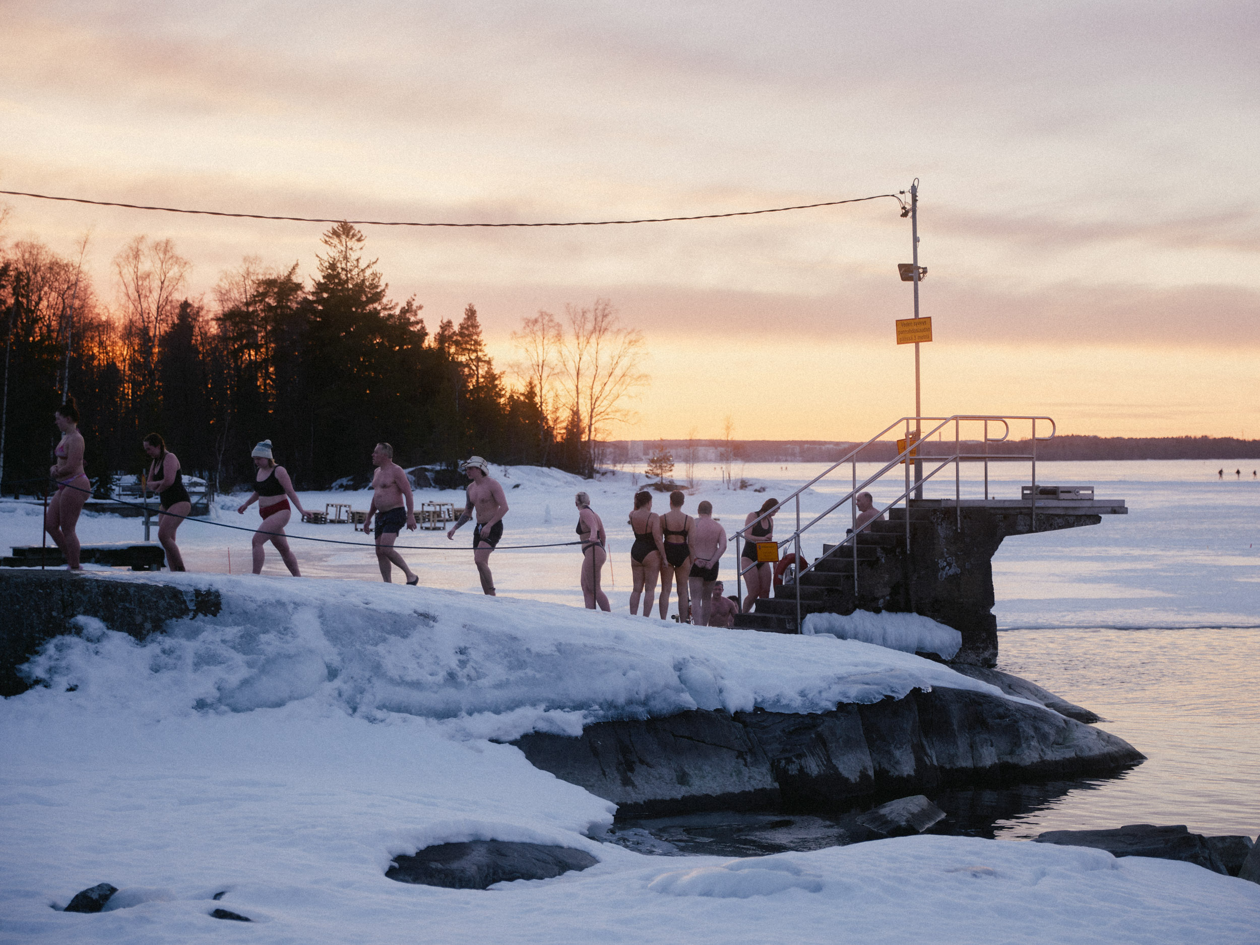 Talviuimareita rivissä jäisellä rannalla auringonlaskun sävyissä.