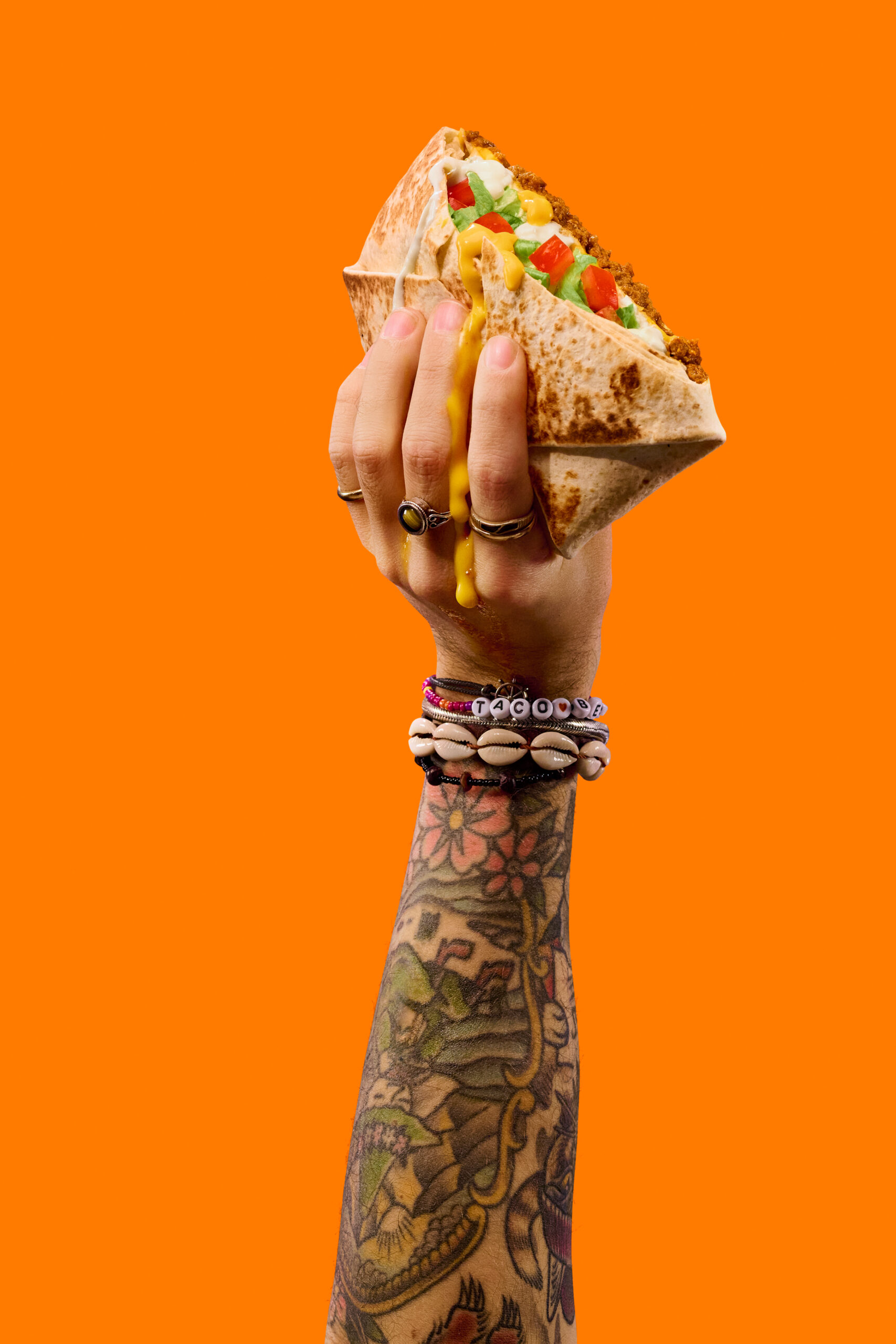 Tatuoitu käsi ja korut pitävät täyteläistä tacoa, jossa on salaattia, tomaattia ja juustoa, oranssin taustan edessä.