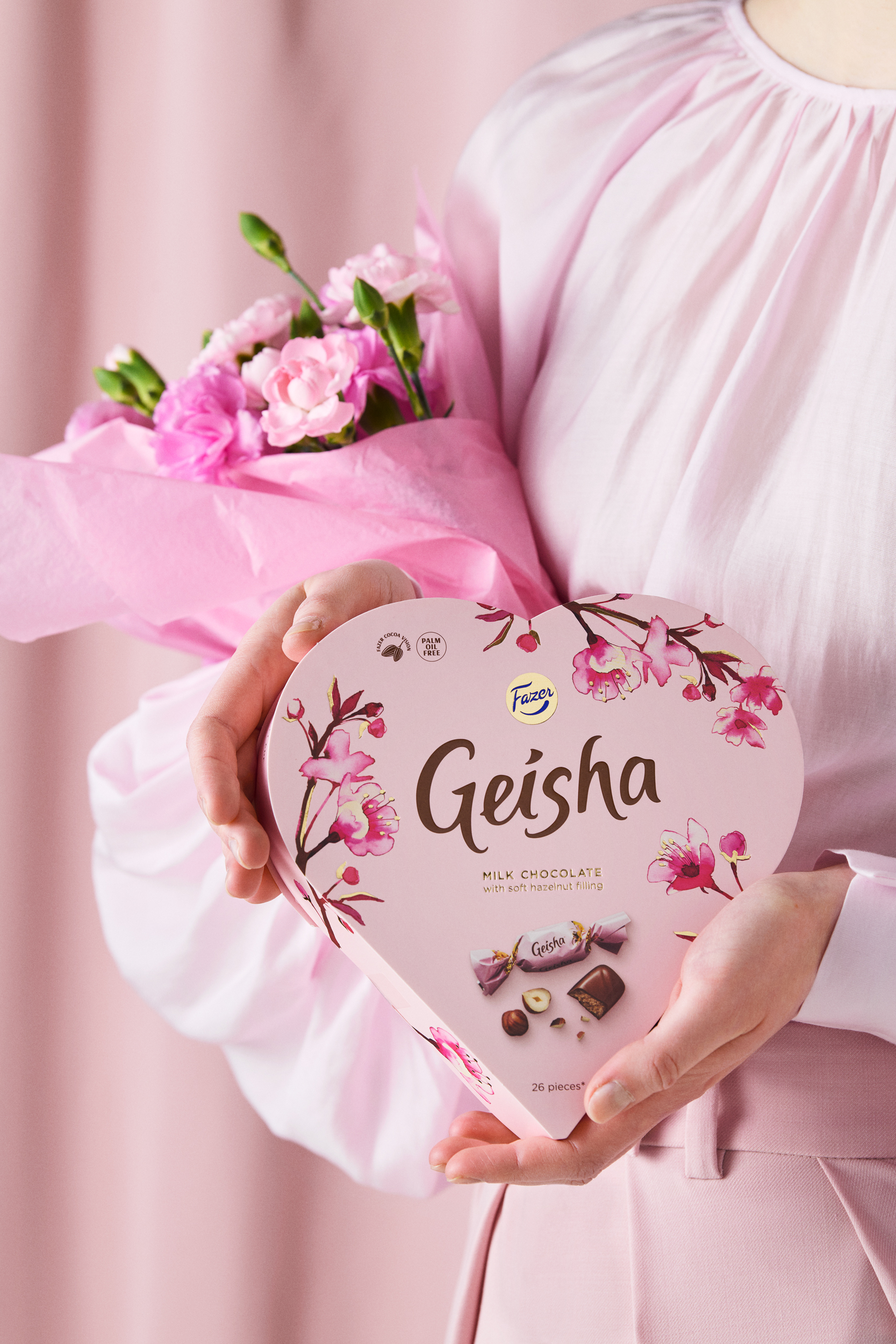 Nainen vaaleanpunaisessa asussa, kädessään sydämenmuotoinen Fazer Geisha-suklaarasia ja pinkki kukkakimppu.
