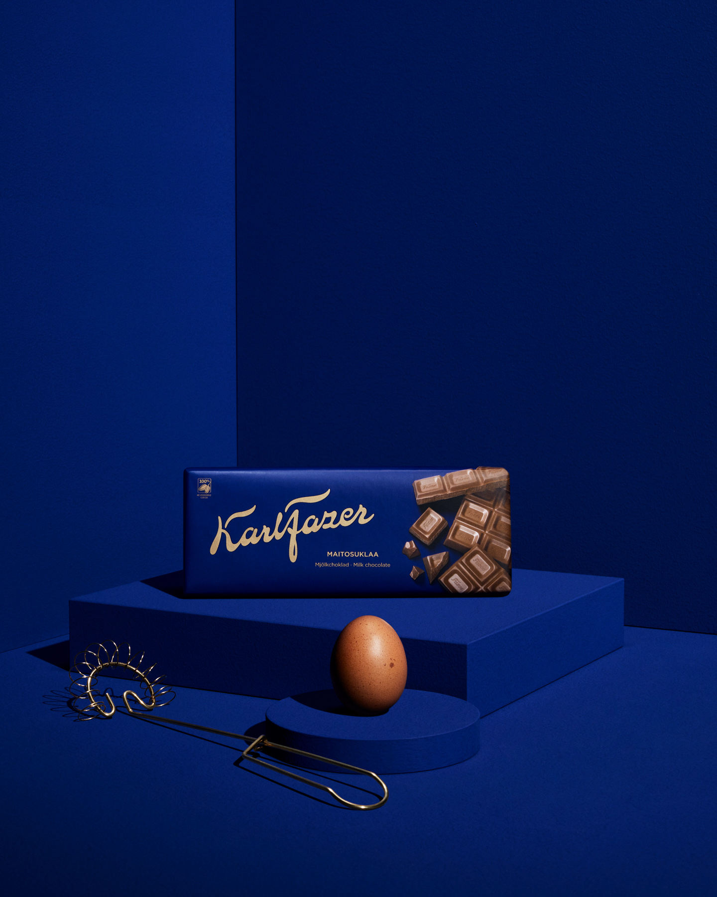 Karl Fazer maitosuklaa -sininen suklaarasia pöydällä, syvä sininen tausta ja koriste-esineet.