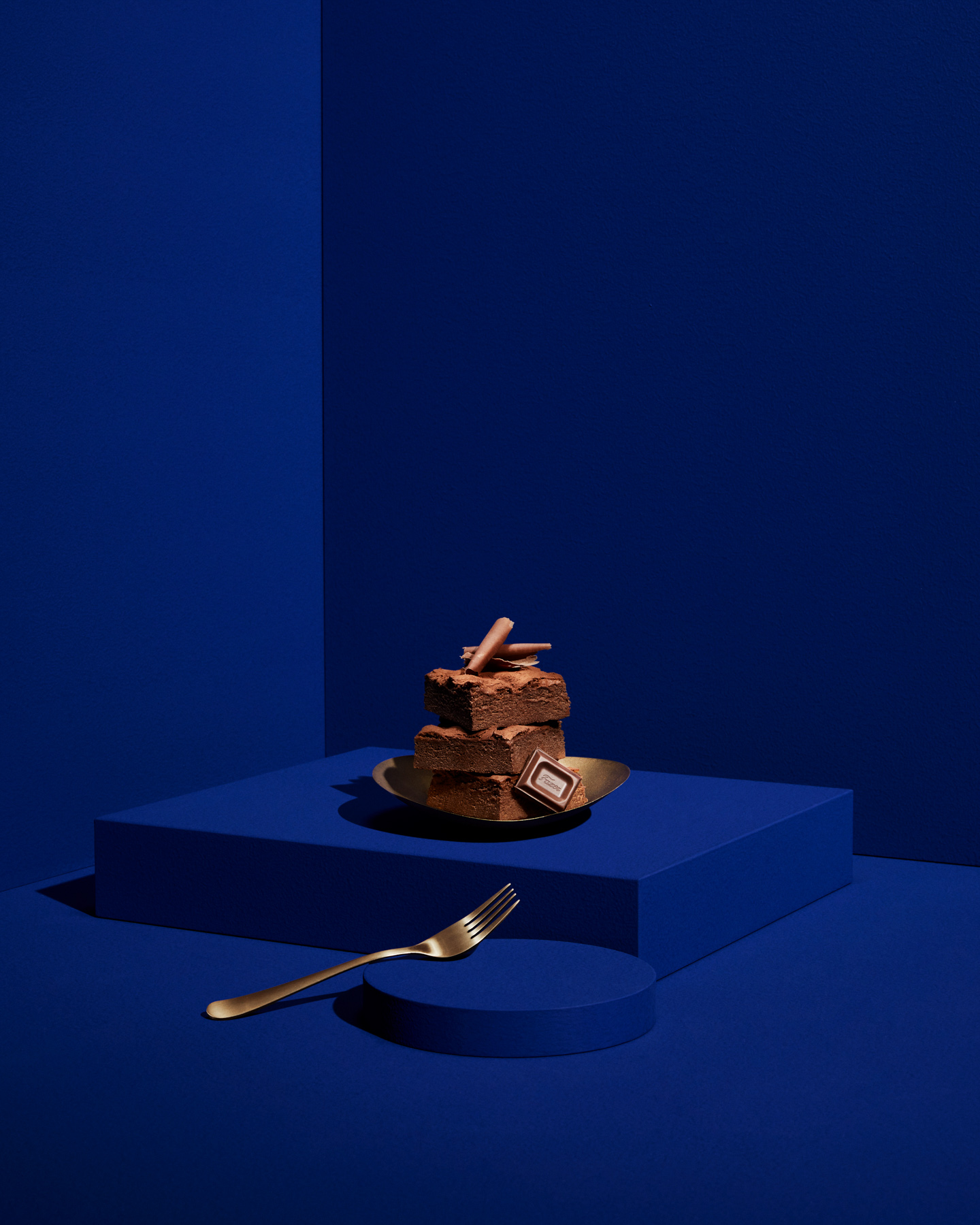 Sinisellä taustalla kerroksittain aseteltu brownie-kakku kultaisella lautasella; suklaapala, kultainen haarukka vasemmalla.