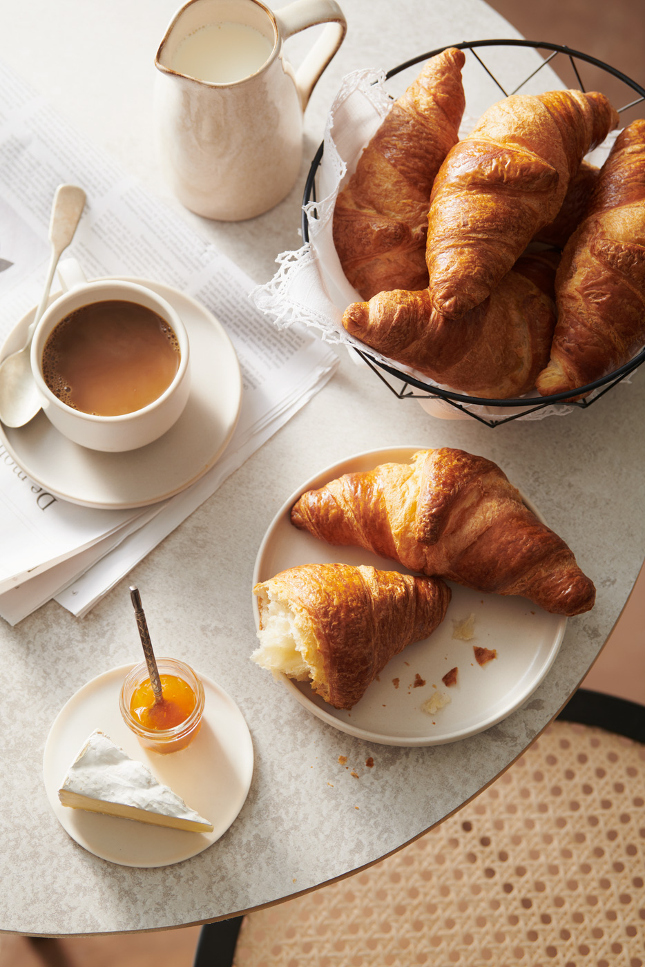 Aamiaispöytä: croissantteja korissa, kuppi kahvia, maitokannu, hillo ja juustoa tarjolla.