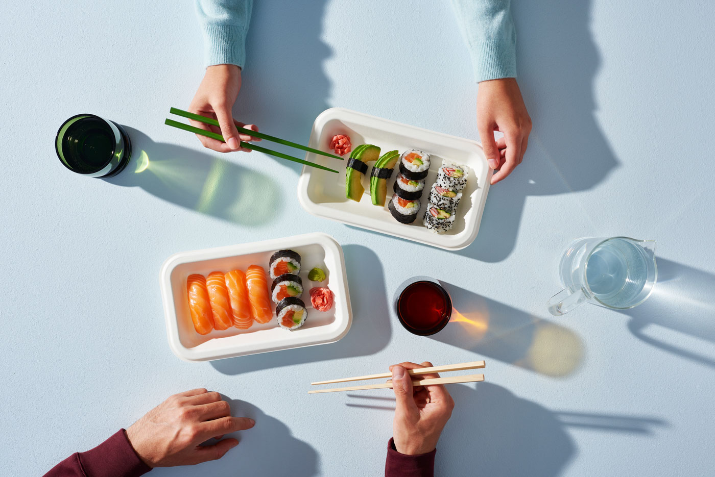 Sushi-lautaita pöydällä: nigiri- ja maki-rullia, wasabi ja inkivääri, sekä kädessä syömäpuikot.