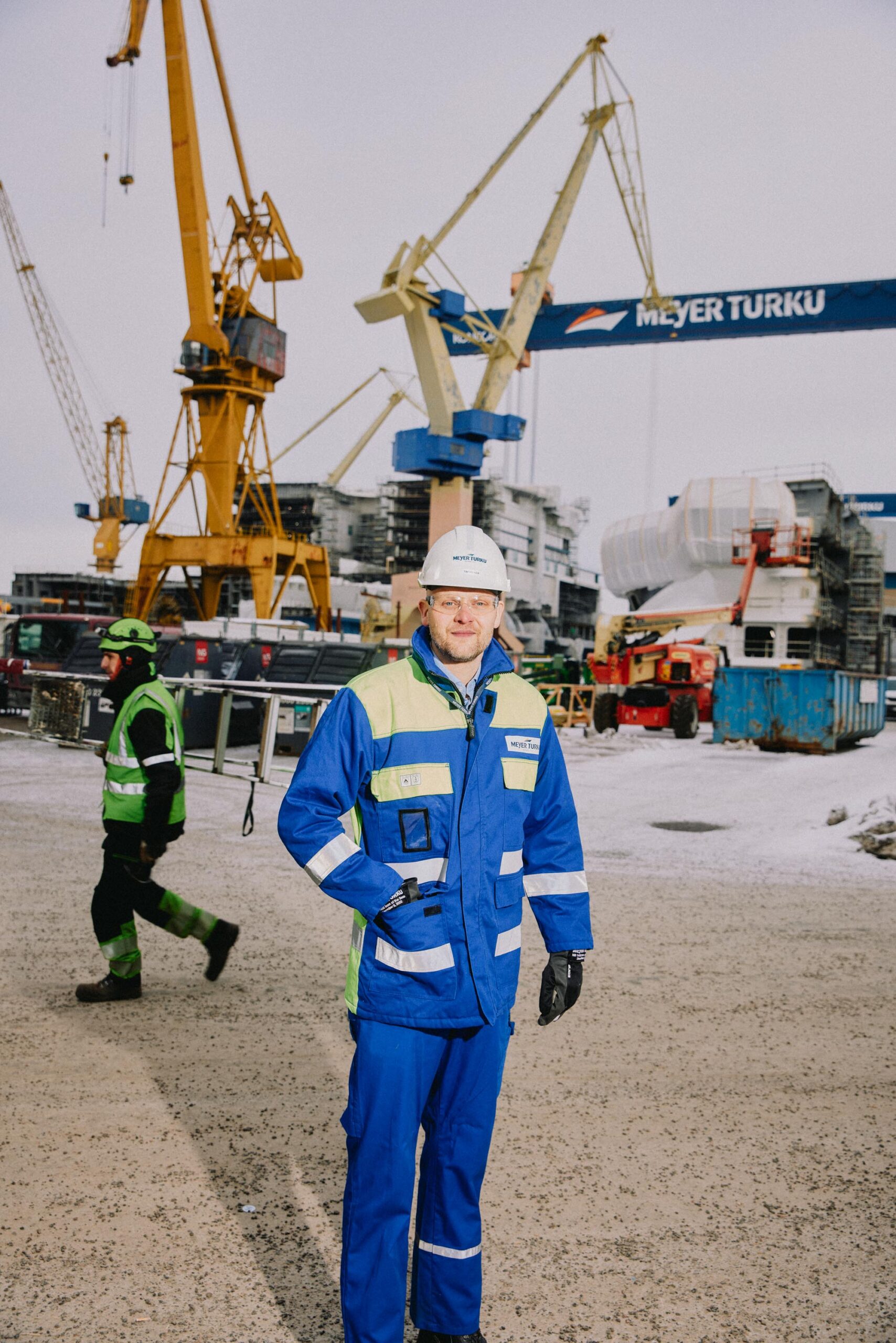 Meyer Turku -rakennustyömaa: työntekijä sinisessä turvaliivissä ja kypärässä etualalla, suuria nostureita taustalla.