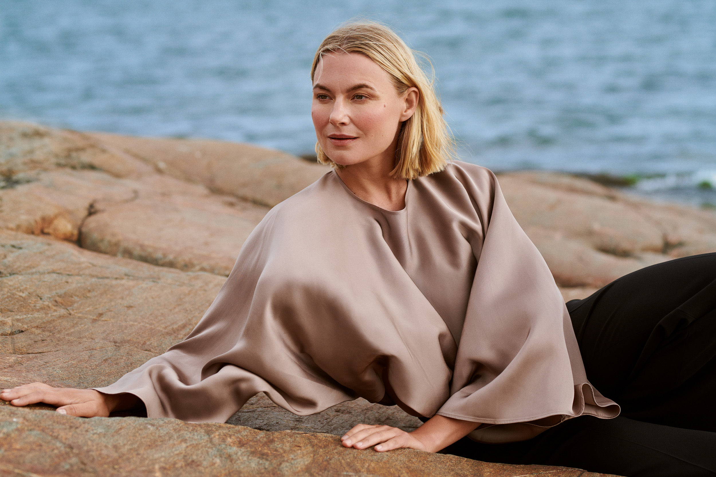 Nainen istuu kalliolla meren rannalla beige silkkisessä topissa, taustalla rauhallinen meri.