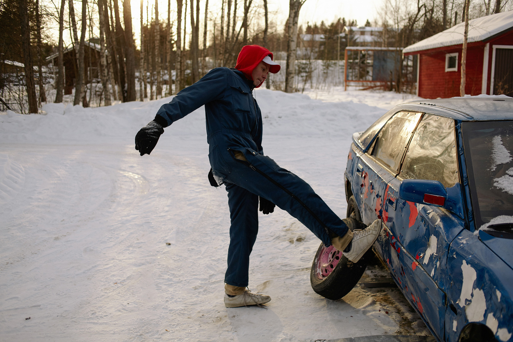 Talvinen pihakuva: mies sinisessä haalarissa potkii vaurioitunutta sinistä autoa lumisella pihalla.