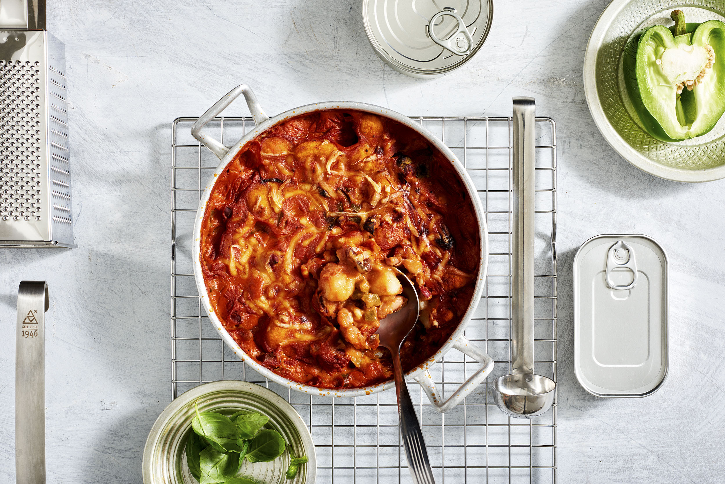 Ylhäältä päin: punainen tomaattikastikkeinen pasta-pata, basilikaa, vihreä paprika ja keittiövälineitä pöydällä.