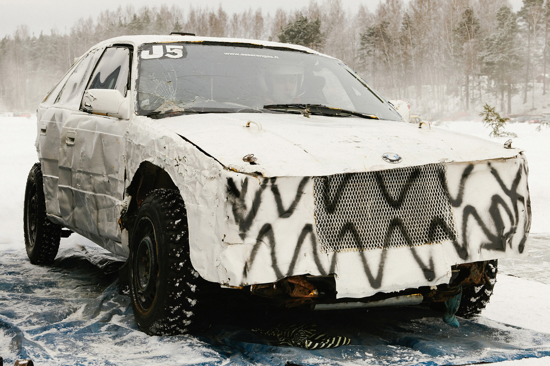 Luminen, graffitilla peitetty ja vaurioitunut auto, rikkoutunut tuulilasi, taustalla talvinen metsä.