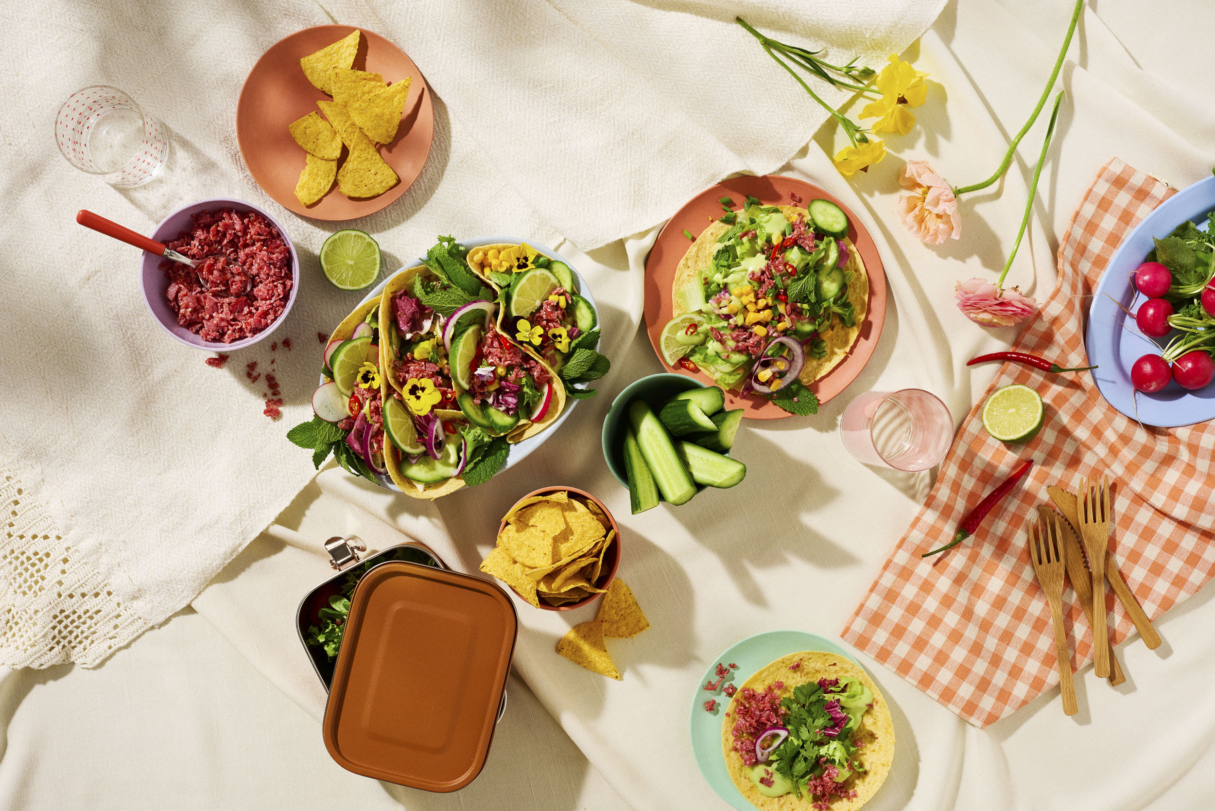 Kesäinen picnic-kattaus tacoilla: täytetyt tacot, nachoja, vihanneksia, lime ja kukkakoristeet pöydällä.