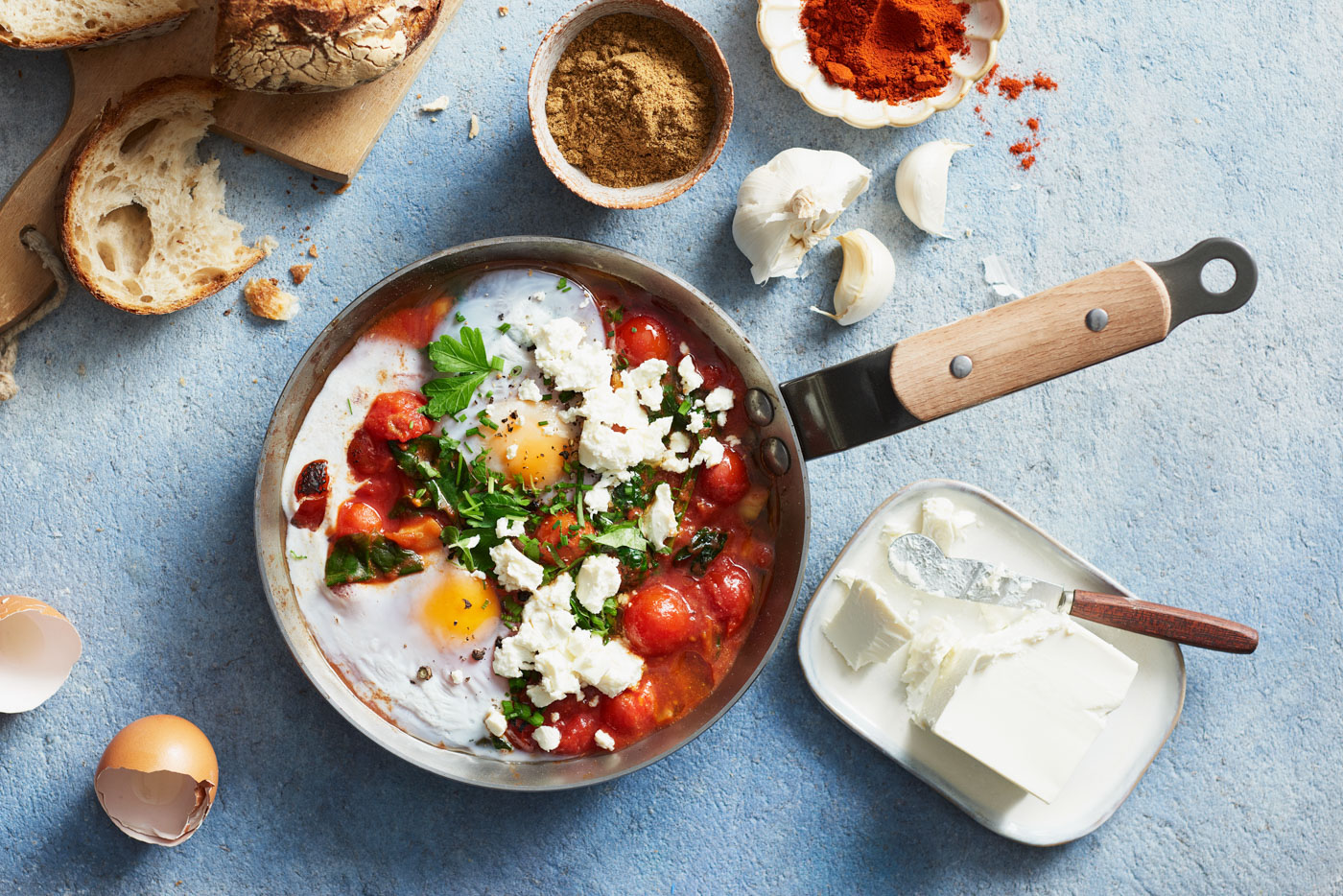 Kananmunat tomaattikastikkeessa pannussa, feta murennettuna ja yrttejä; leipä ja mausteet pöydällä.