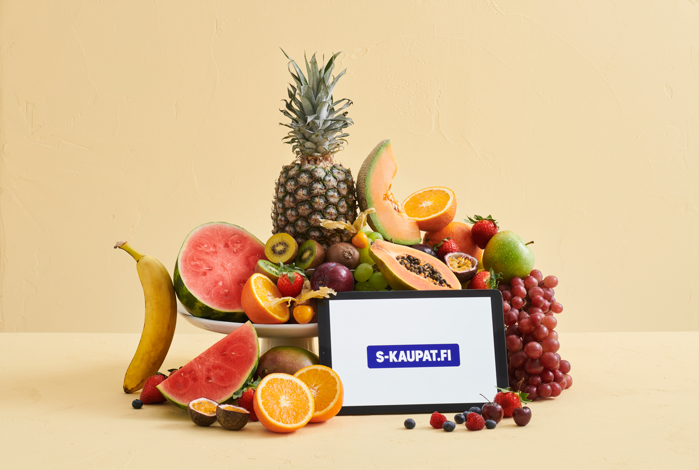Runsas hedelmäkori, ananas, meloni, appelsiinit ja marjat sekä tabletilla lukee S-KAUPAT.FI.