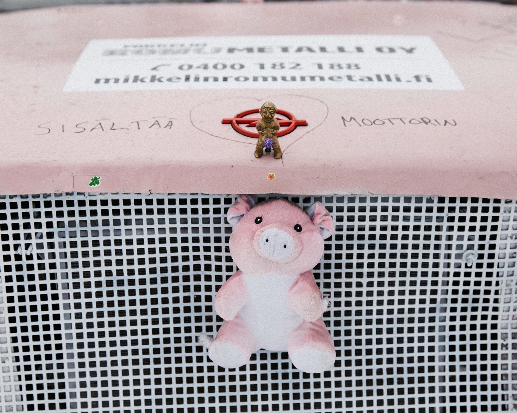 Pieni muovihahmo seisoo punaisen ympyrän keskellä pinkillä pöydällä; taustalla ritilä ja alhaalla pinkki pehmopossu.
