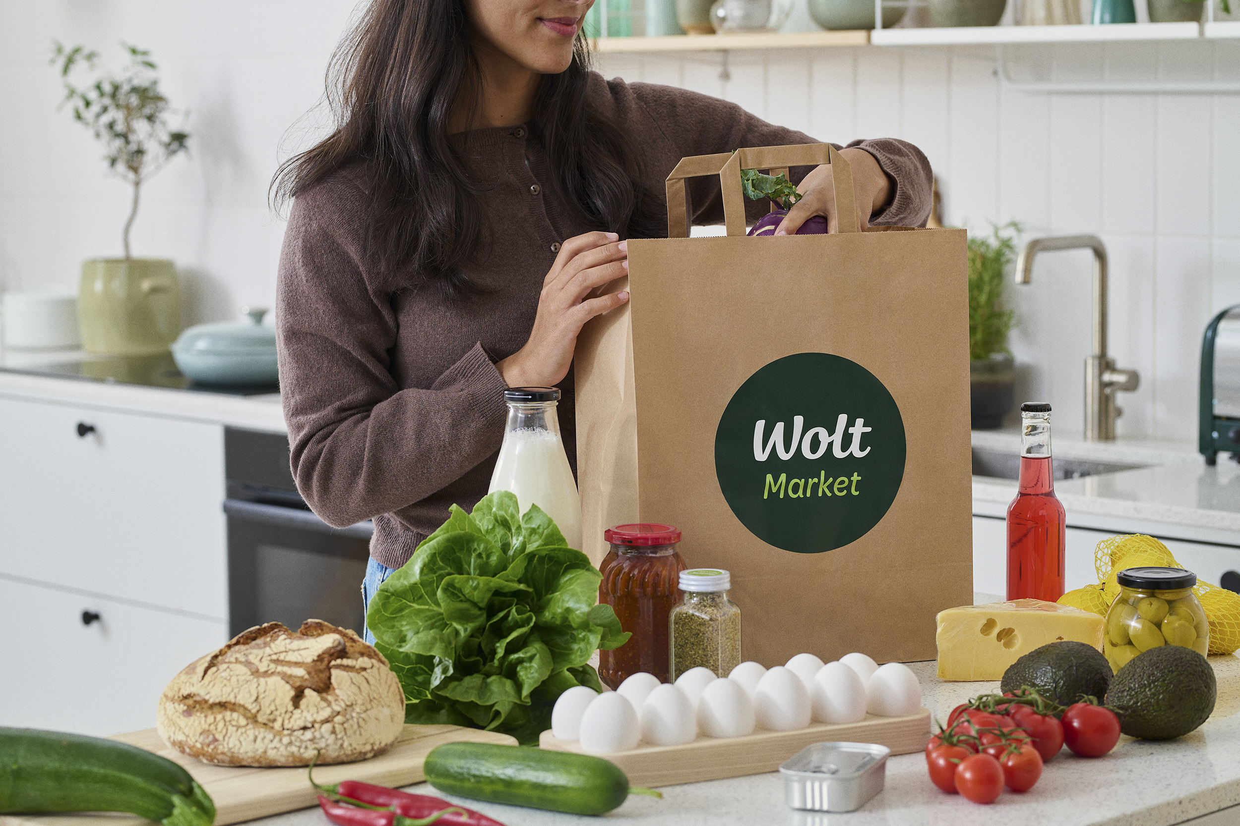 Nainen pakkaa Wolt Market -ostoskassin keittiössä tuoreita vihanneksia, leipää ja maitotuotteita.