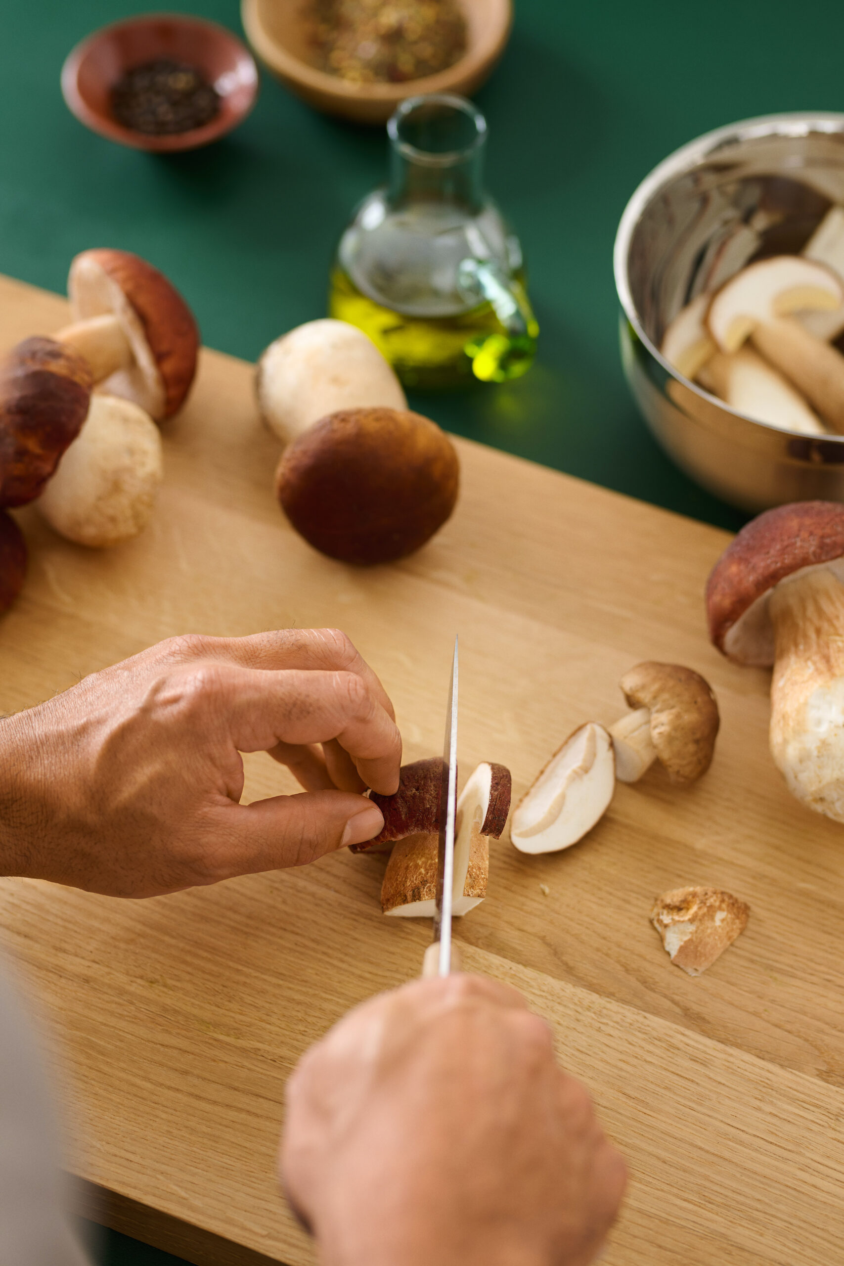 Kädet leikkaamassa sieniä pöydällä; taustalla oliiviöljypullo ja kulhoja sekä leipäpalasia.