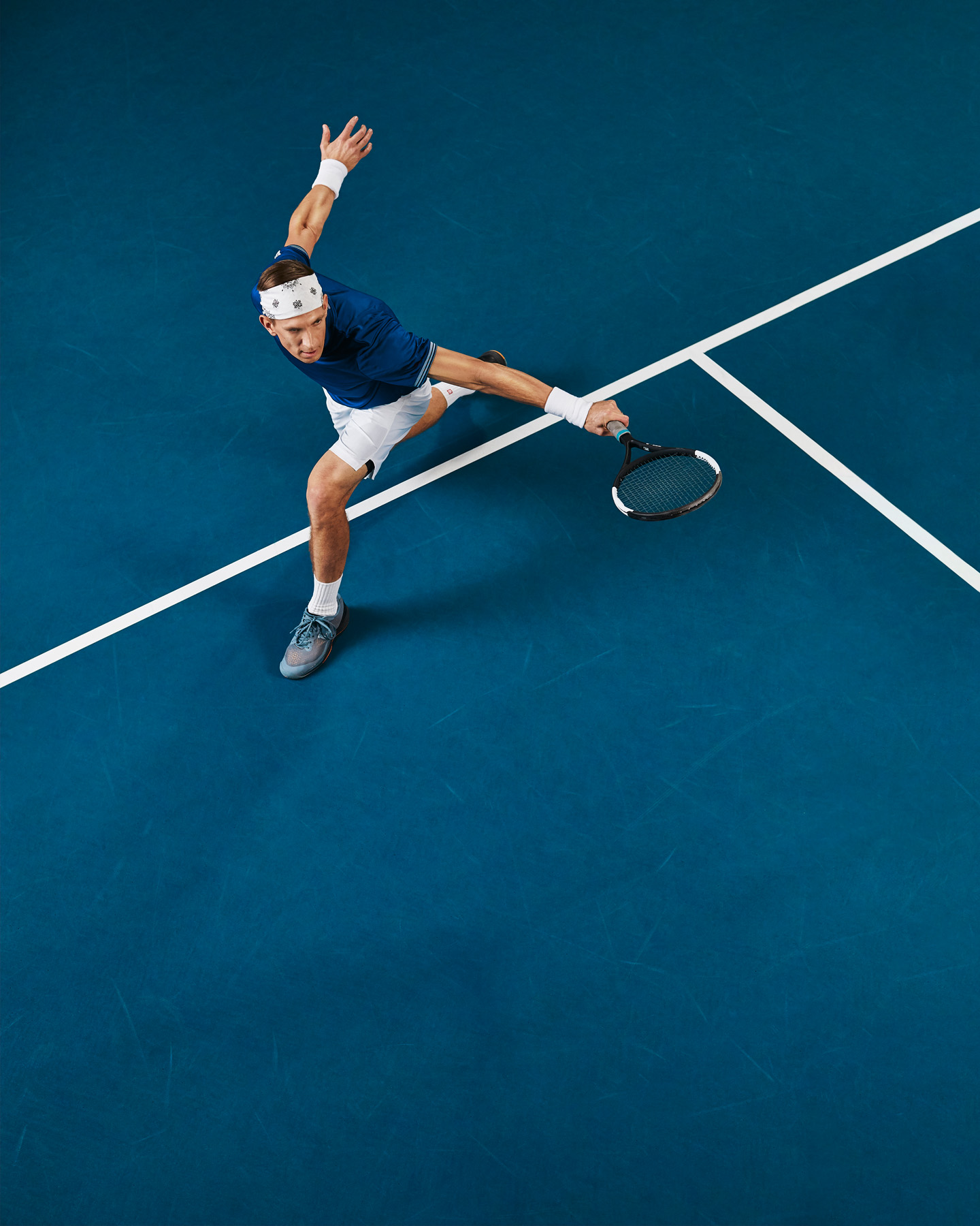 Tennispelaaja iskee forehand-lyöntiä sinisellä tenniskentällä ylhäältä; sininen paita, valkoiset shortsit ja päänauha.