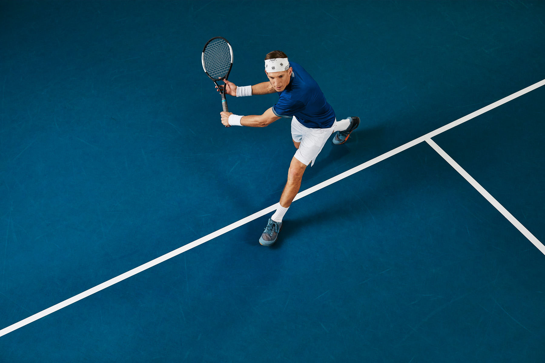 Tennispelaaja sinisellä kentällä ylhäältä päin kuvattuna lyömässä palloa.