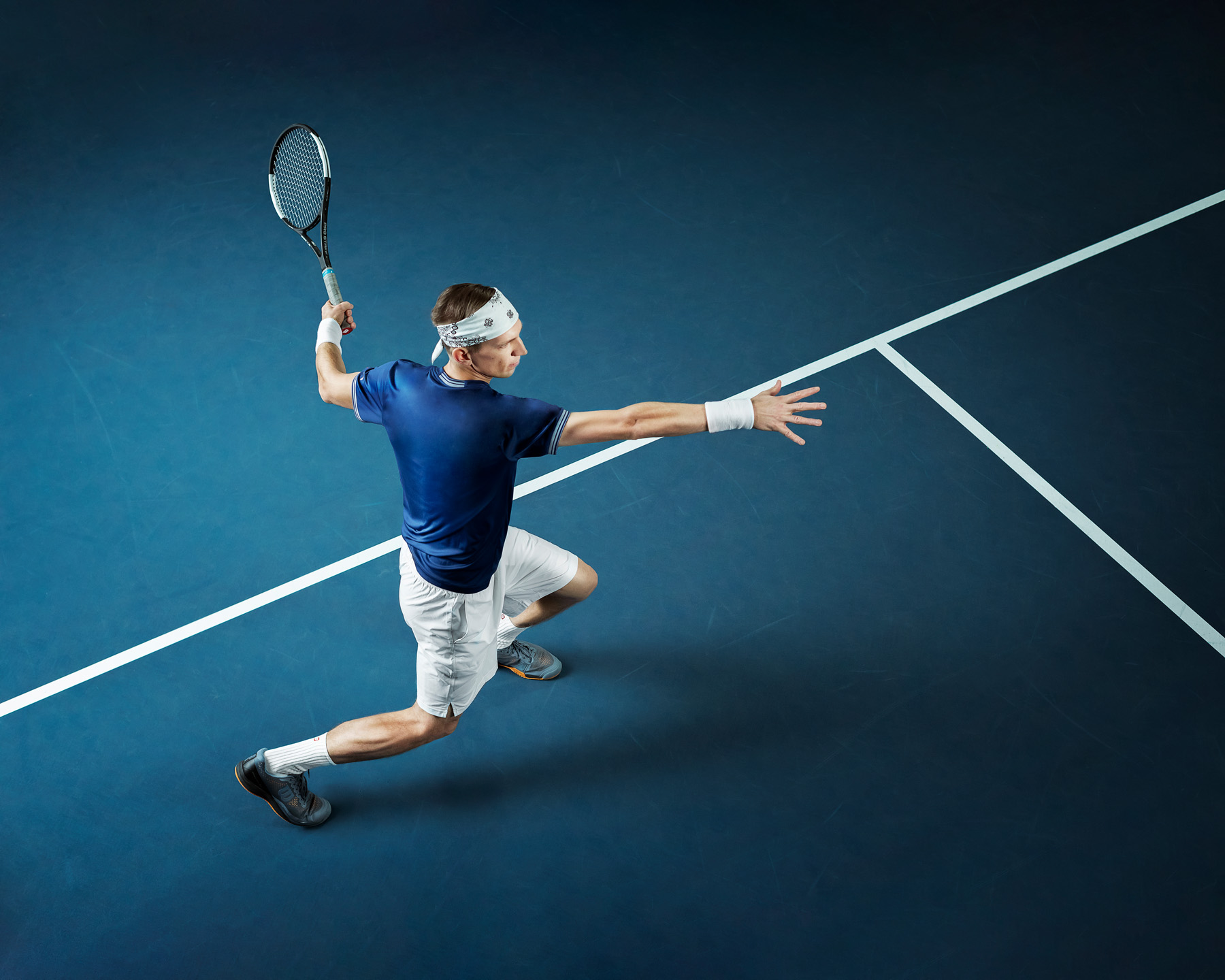 Tennispelaaja hyppää ilmassa lyömässä palloa sinisellä tenniskentällä, päässä valkoinen päänauha.