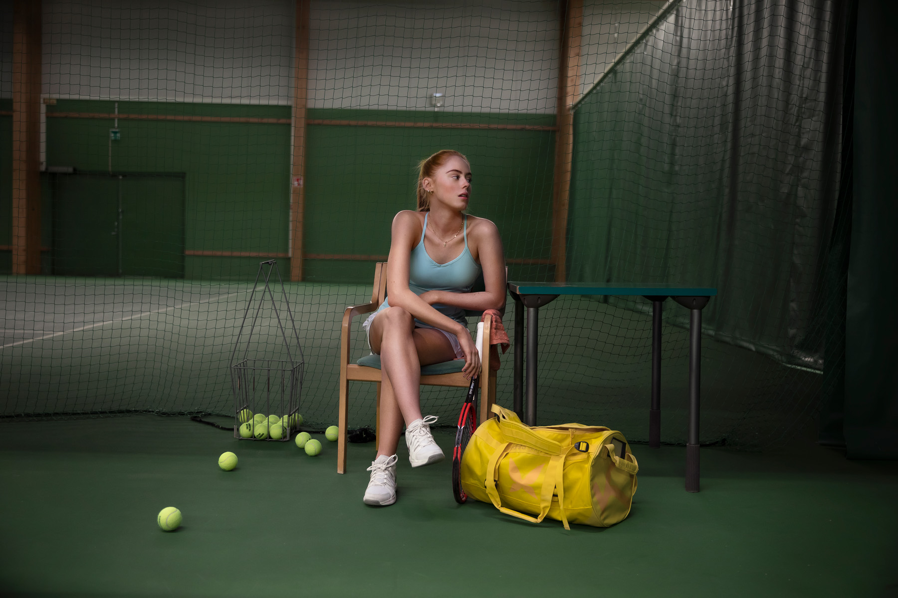 Sisätiloissa tenniskentällä istuva nainen treenivarusteineen, ympärillä maila, kassi ja tennispalloja.