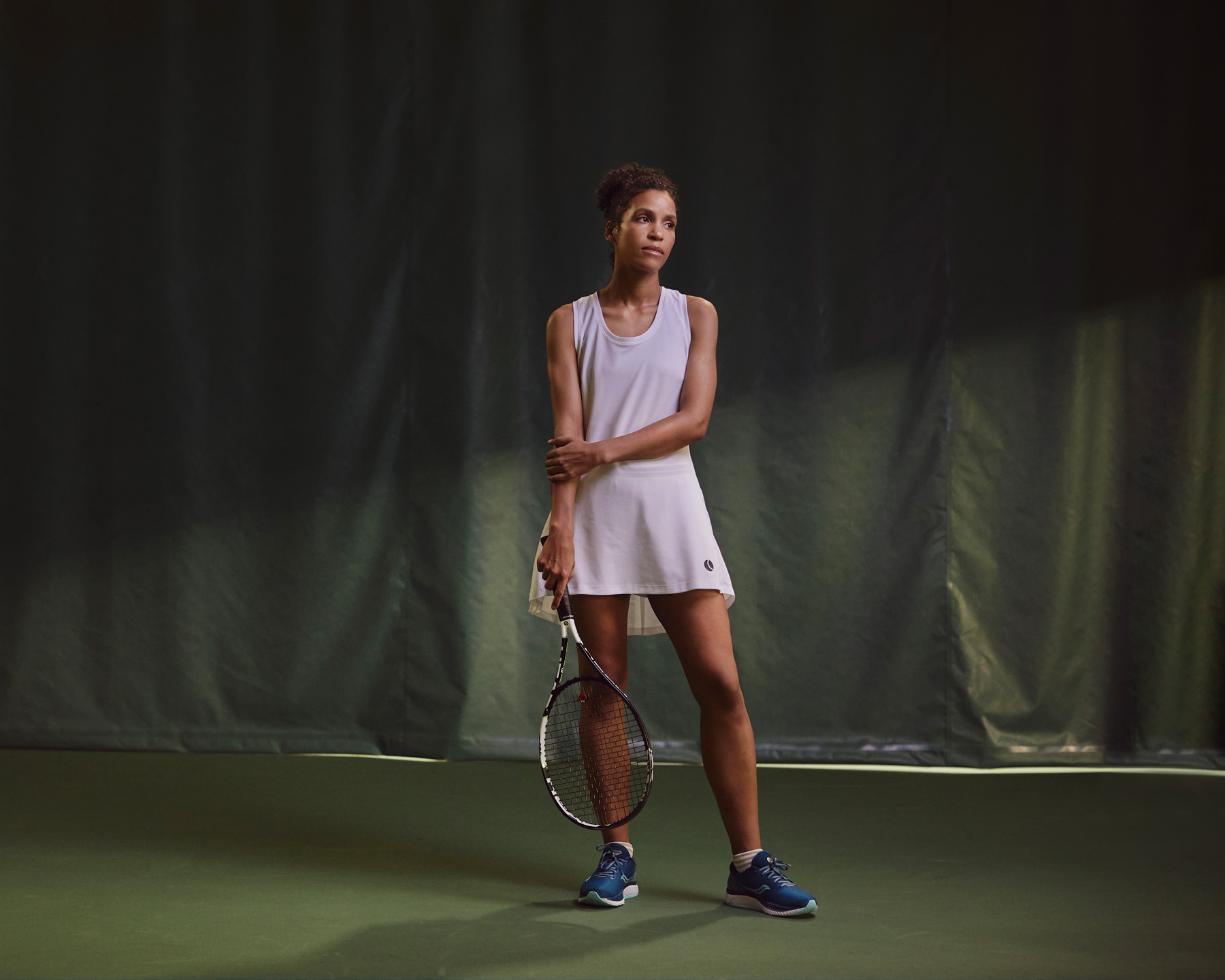 Tennispelaaja nainen valkoisessa treenimekossa seisoo tenniskentällä maila kädessä tummaa taustakangasta vasten.