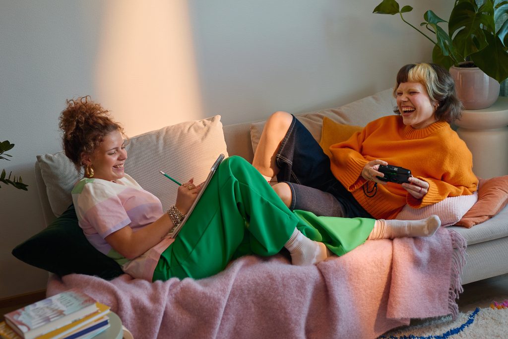 Kaksi nauravaa naista viettää rentoa hetkeä sohvalla. Toinen kirjoittaa muistilehtiöön ja toinen pitelee kameraa käsissään.